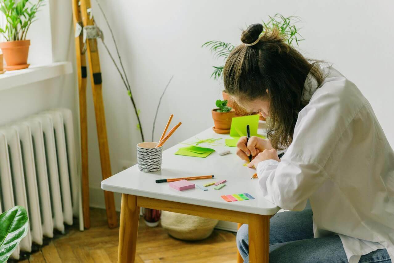 A young woman writes on sticky notes at a desk.