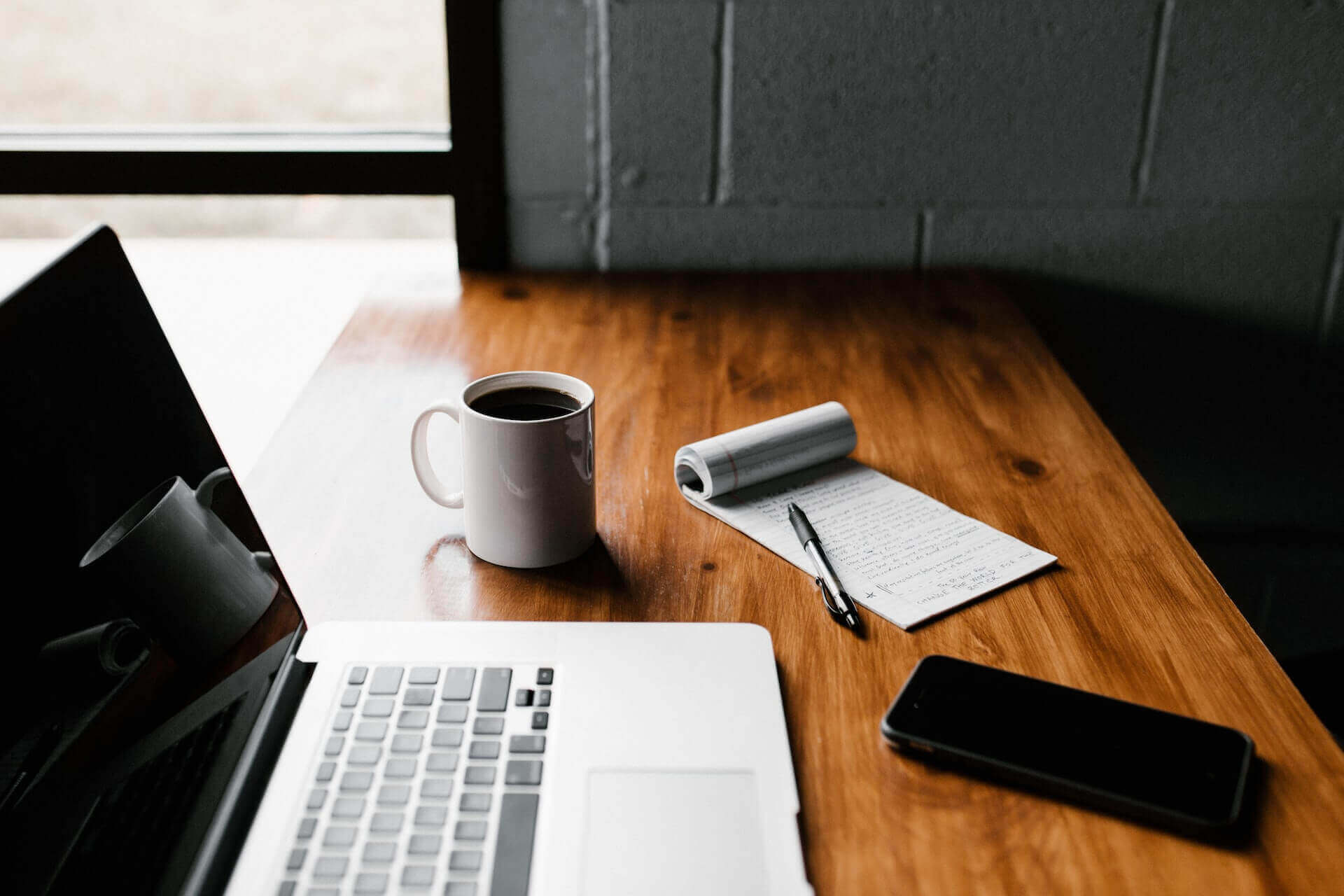 A notepad with writing lays on a table next to a smartphone, laptop, and coffee mug.