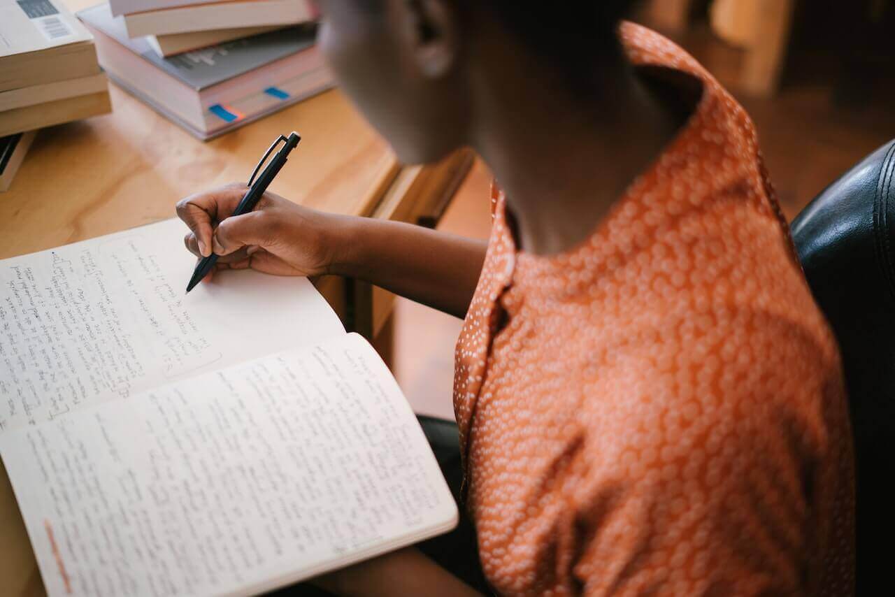A young adult writes in a notebook, with textbooks nearby.